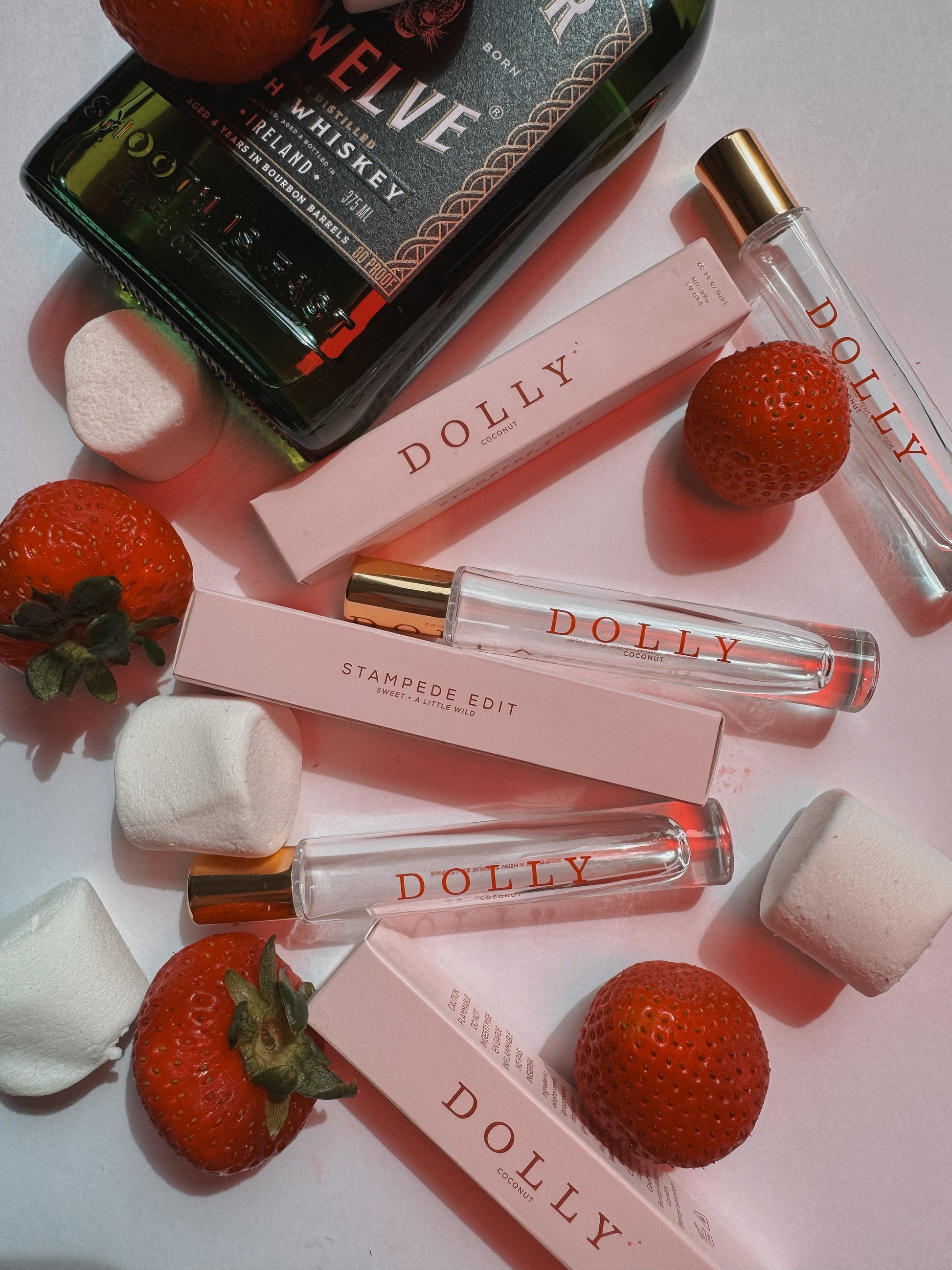 Dolly perfume travel atomizers and their pink boxes arranged on a pink surface with strawberries and marshmallows, alongside a bottle of whiskey as a styled prop.