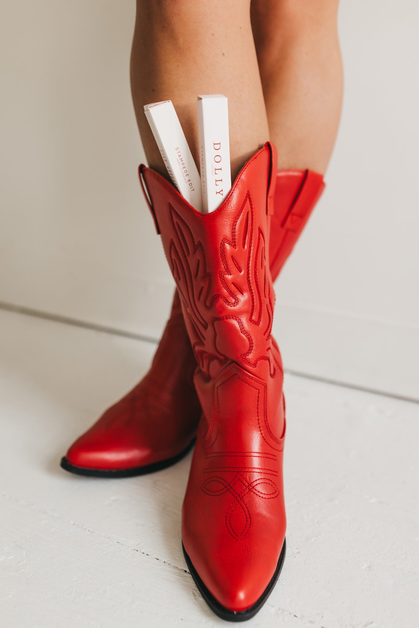 A pair of bright red leather cowboy boots with decorative cutout stitching, shown on a model's feet against a clean white background. Two white books with 'BOHLY' printed on them are tucked into the shaft of the front boot.
