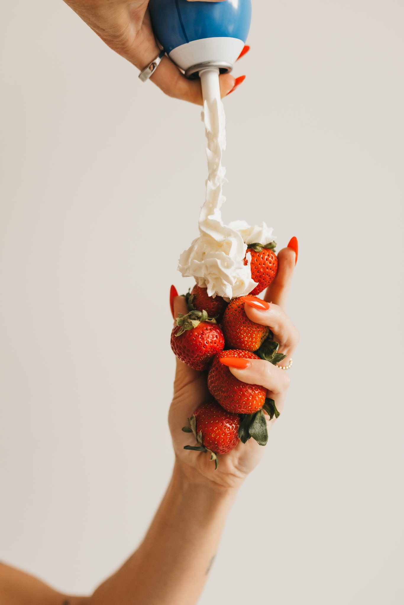 A close-up of a hand holding a bunch of fresh red strawberries while another hand dispenses whipped cream from a blue and white can onto the strawberries against a plain light background.